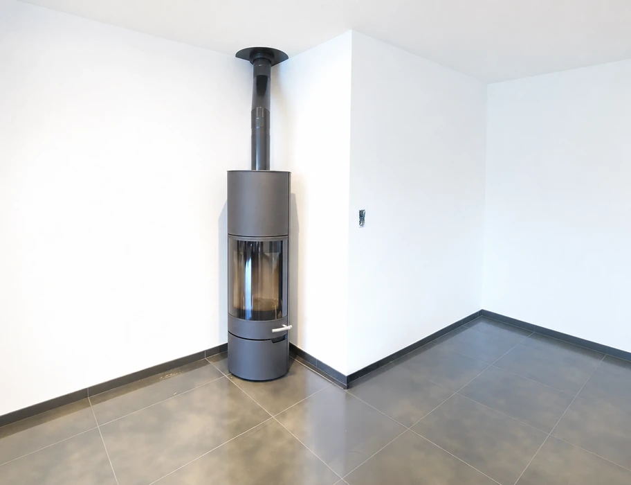 Poêle à bois design cylindrique de style contemporain, en acier émaillé anthracite, installé dans un angle de pièce aux murs blancs et sol en carrelage gris foncé